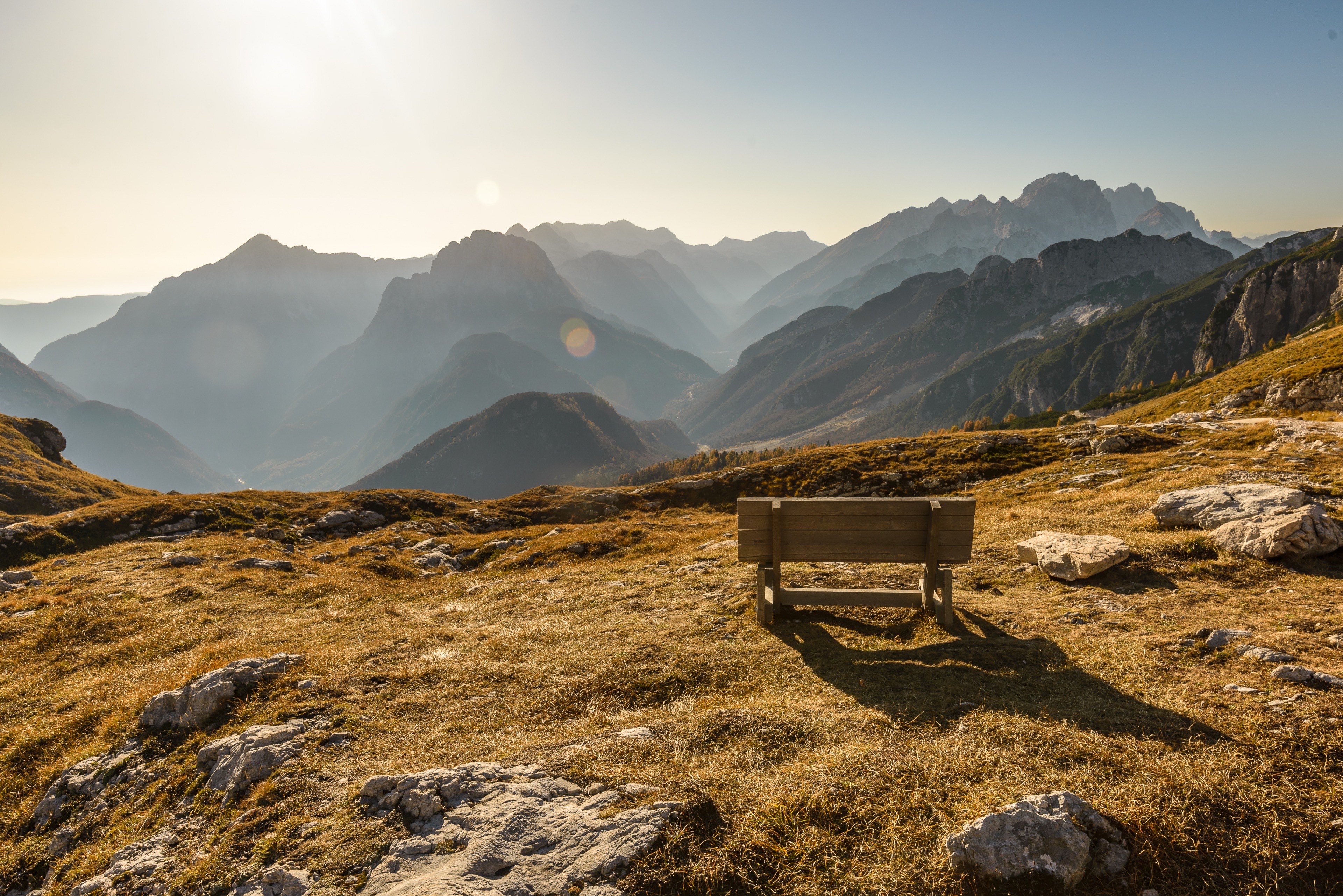 Leere Holzbank auf einer goldenen Alpwiese mit Blick über gestaffelte Berge im Abendlicht