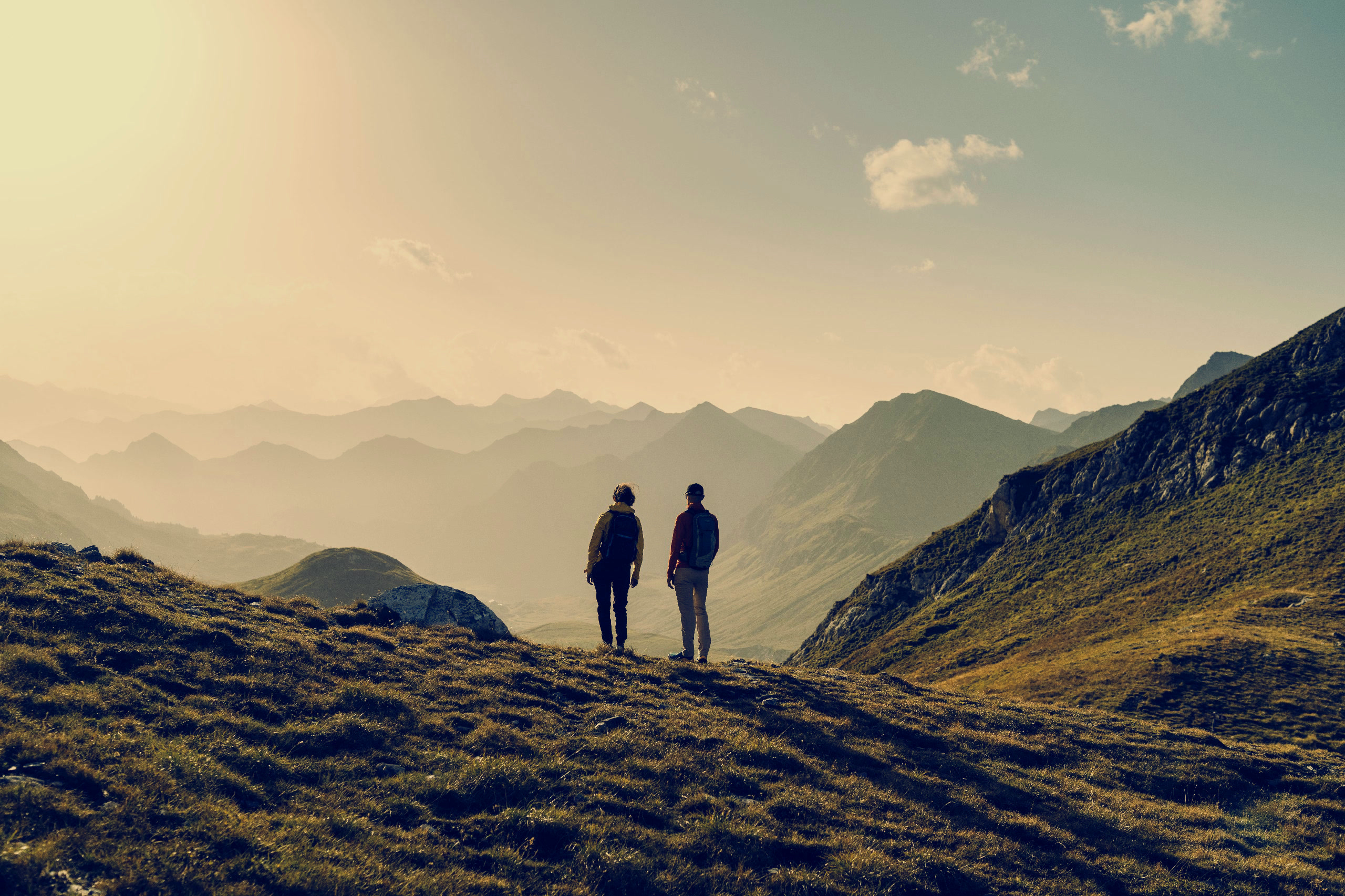 Zwei Wanderer auf einem Alpengrat im goldenen Abendlicht mit Blick über gestaffelte Schweizer Berge